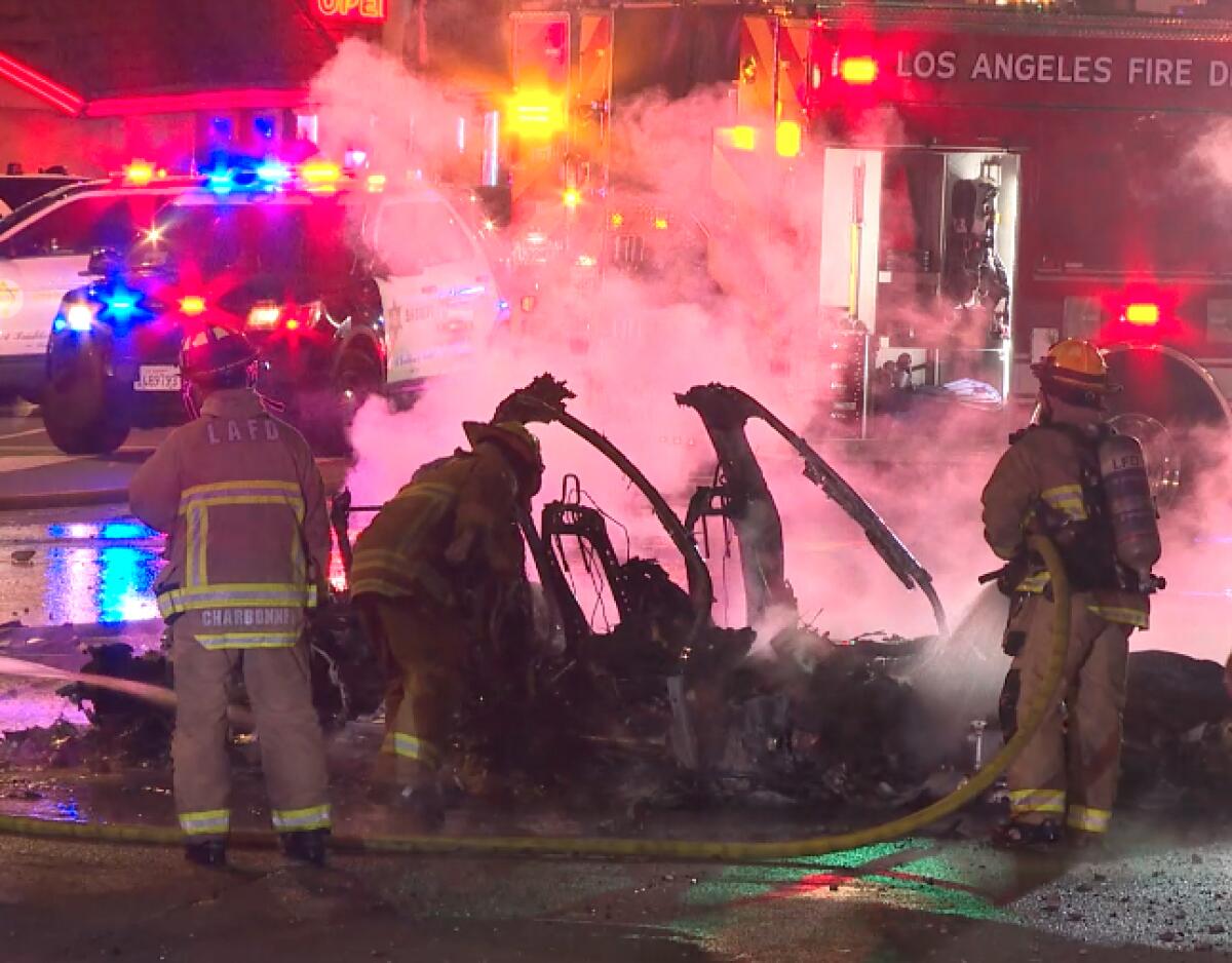Firefighters spray water onto the smoldering shell of a car in the street