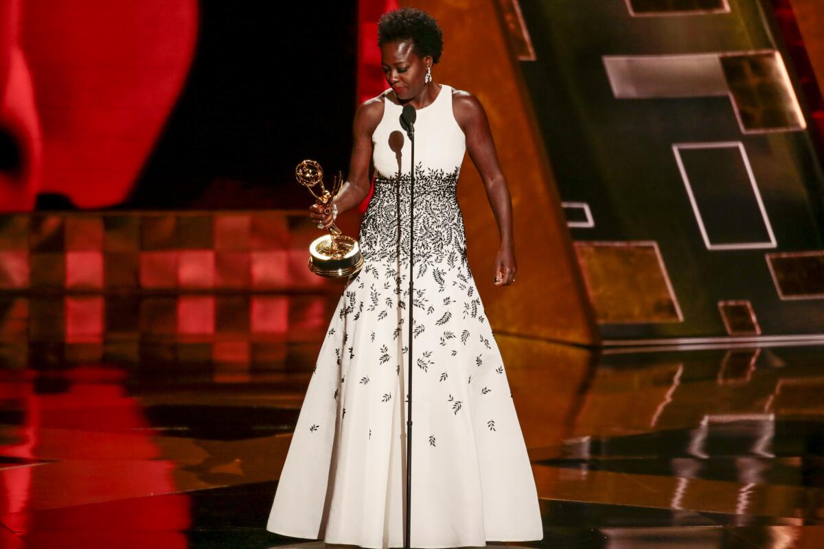 A woman in a long white and black dress stands onstage holding an Emmy Award.