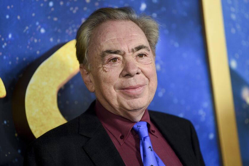 Andrew Lloyd Webber smiling in a black suit, maroon collared shirt and purple tie against a blue and gold background