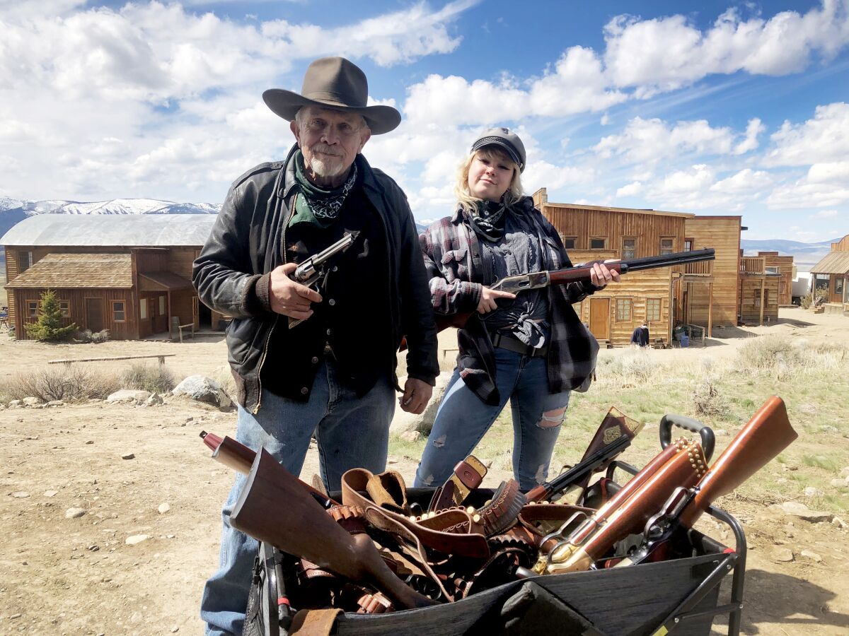 An older man and his daughter stand on an outdoor film set, each holding a gun, with more guns in bins in front of them.