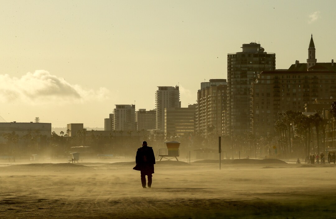 March 9: A person walks on sand as gusty winds raise clouds of dust on a beach with buildings behind it.