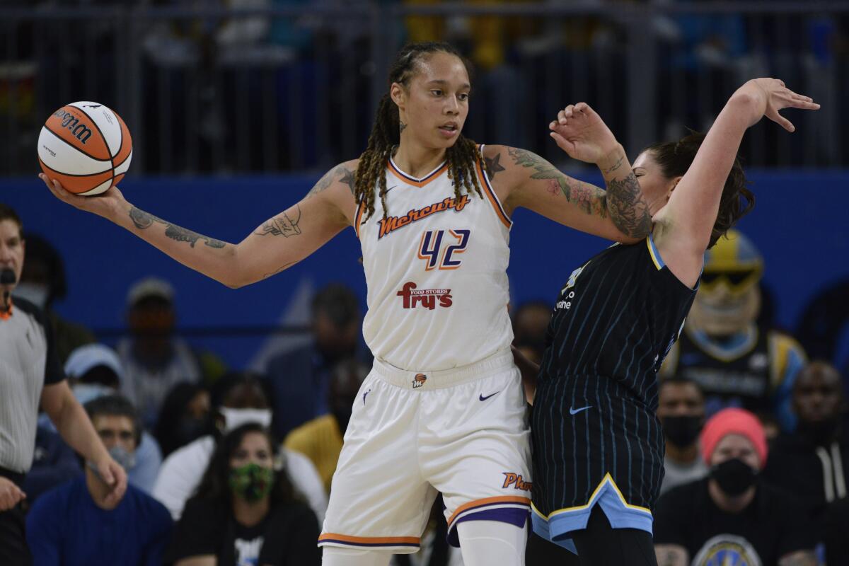 A basketball player holds the ball in her outstretched right arm while fending off a defender with her left elbow