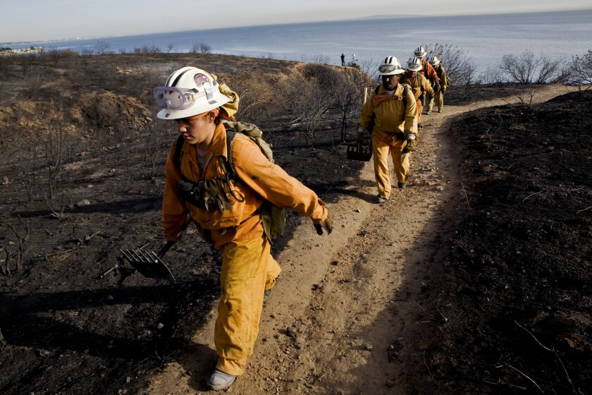 Women in fire suits and helmets walk along a trail with the ocean in the background