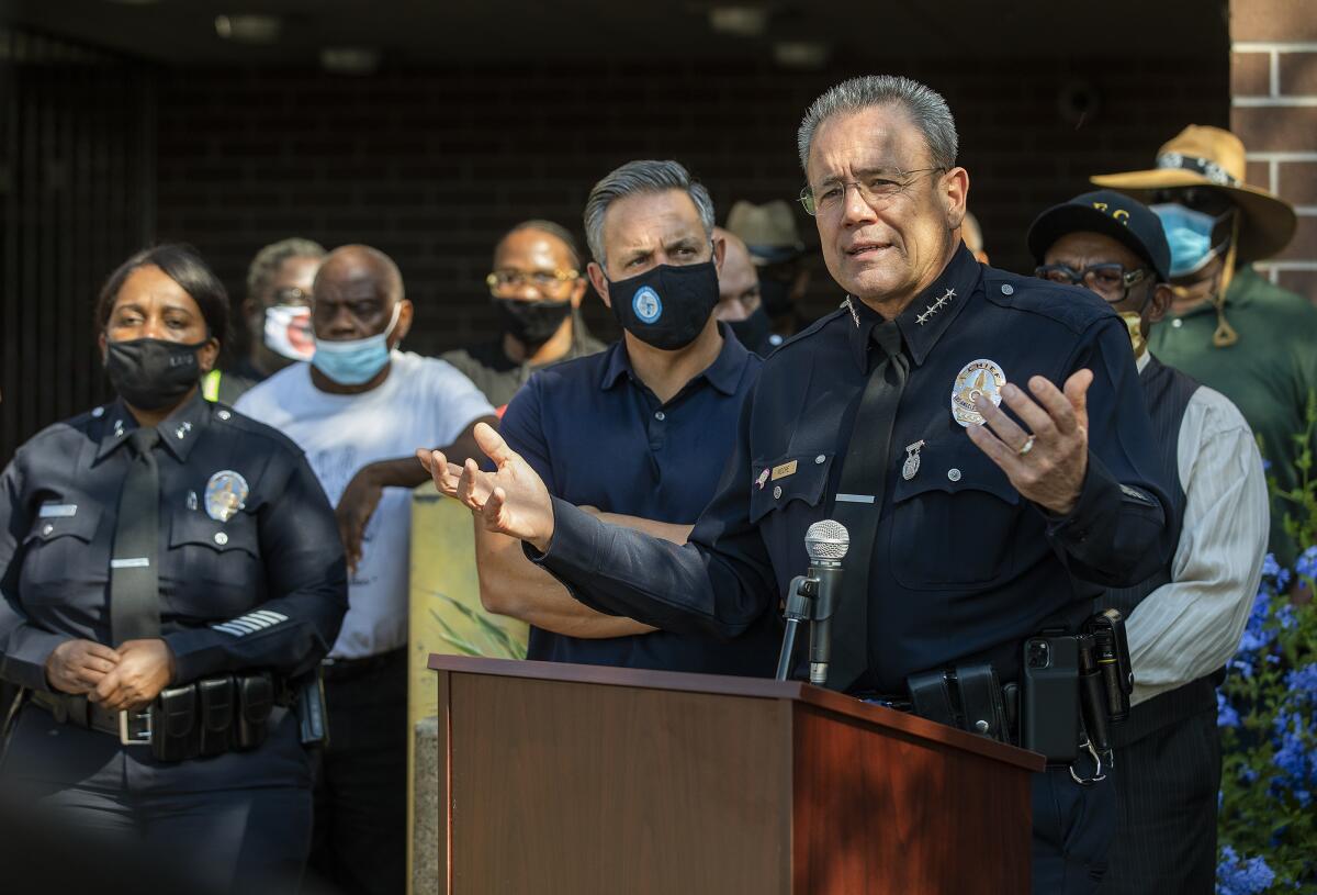 LAPD Chief Michel Moore speaks at a lectern while people stand behind him