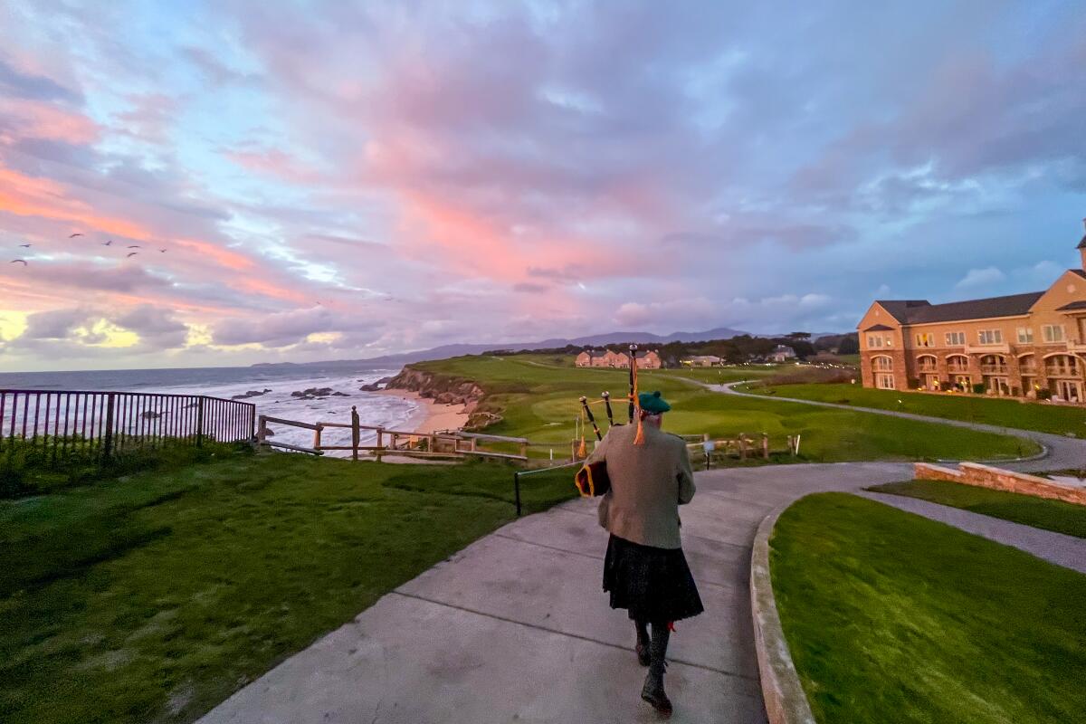 A bagpiper walks along a paved trail near a bay with a beautiful sunset in front.