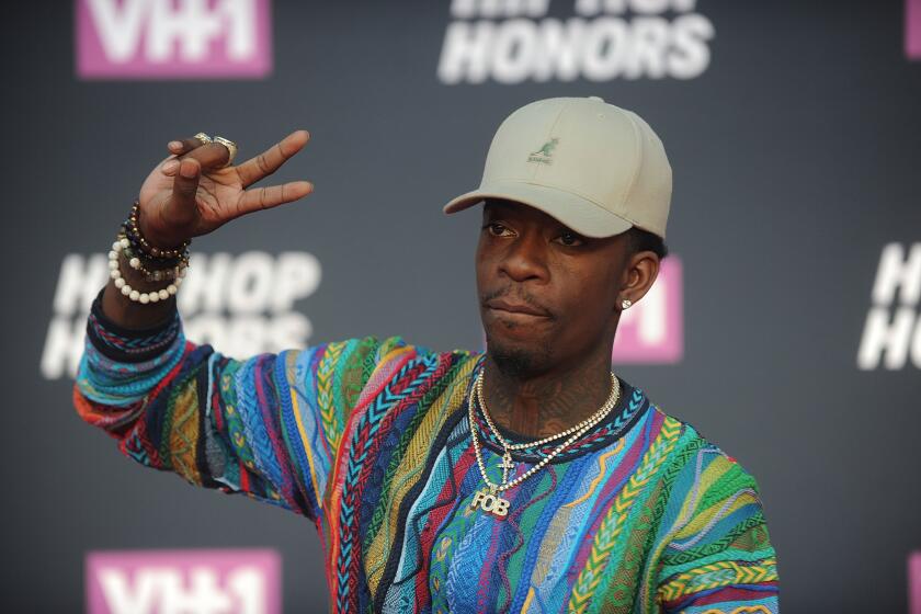 Rich Homie Quan in a muted green hat and a colorful sweater holding up a peace sign and posing in front of a black background