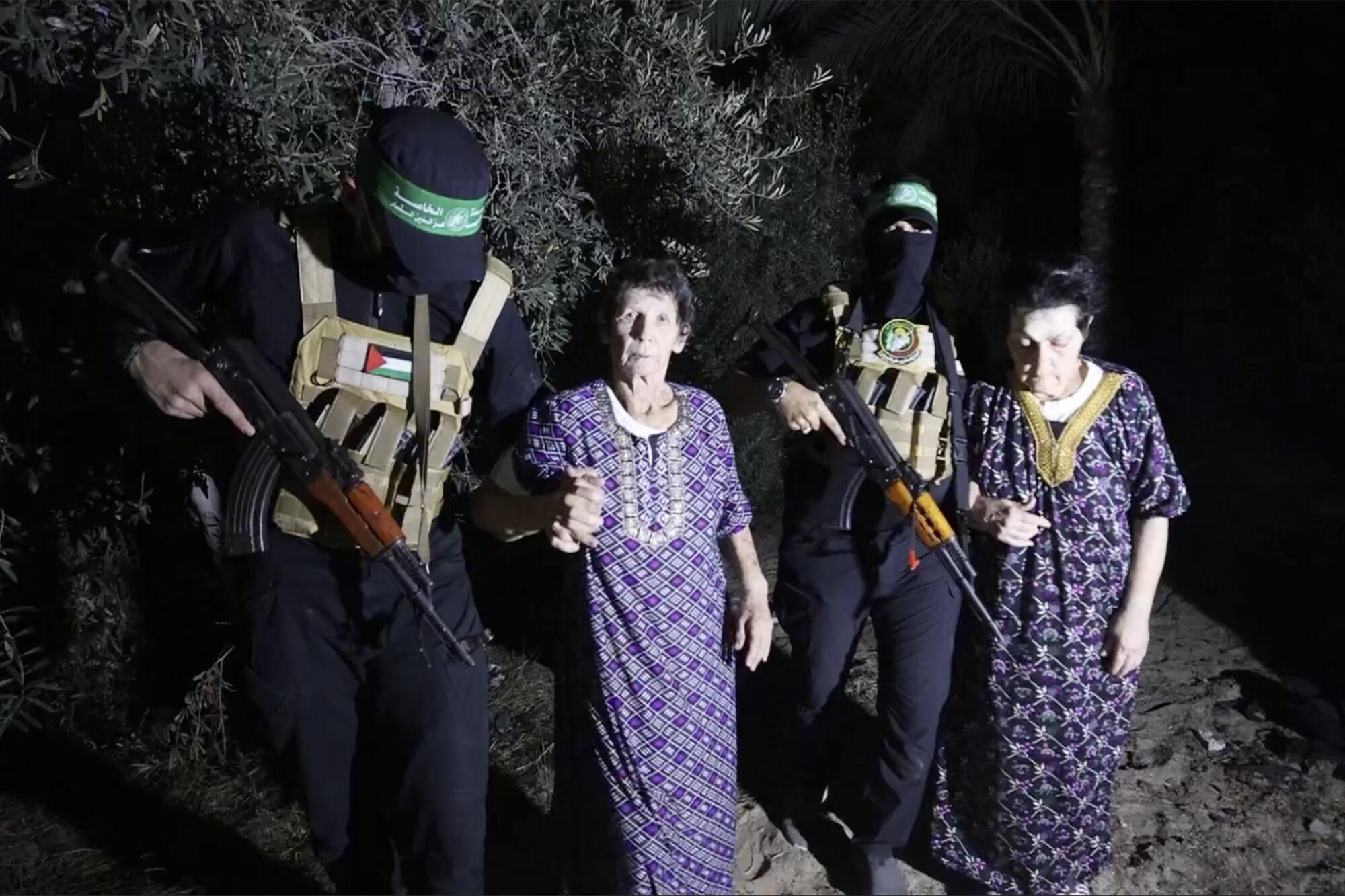 Two masked gunmen in black next to two women in long, printed dresses.