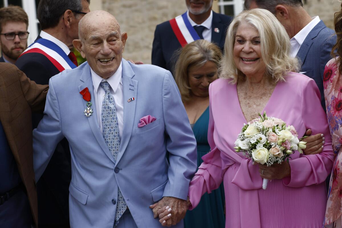 An elderly man in light blue suit and woman in bright pink holding a bouquet clasp hands and smile.
