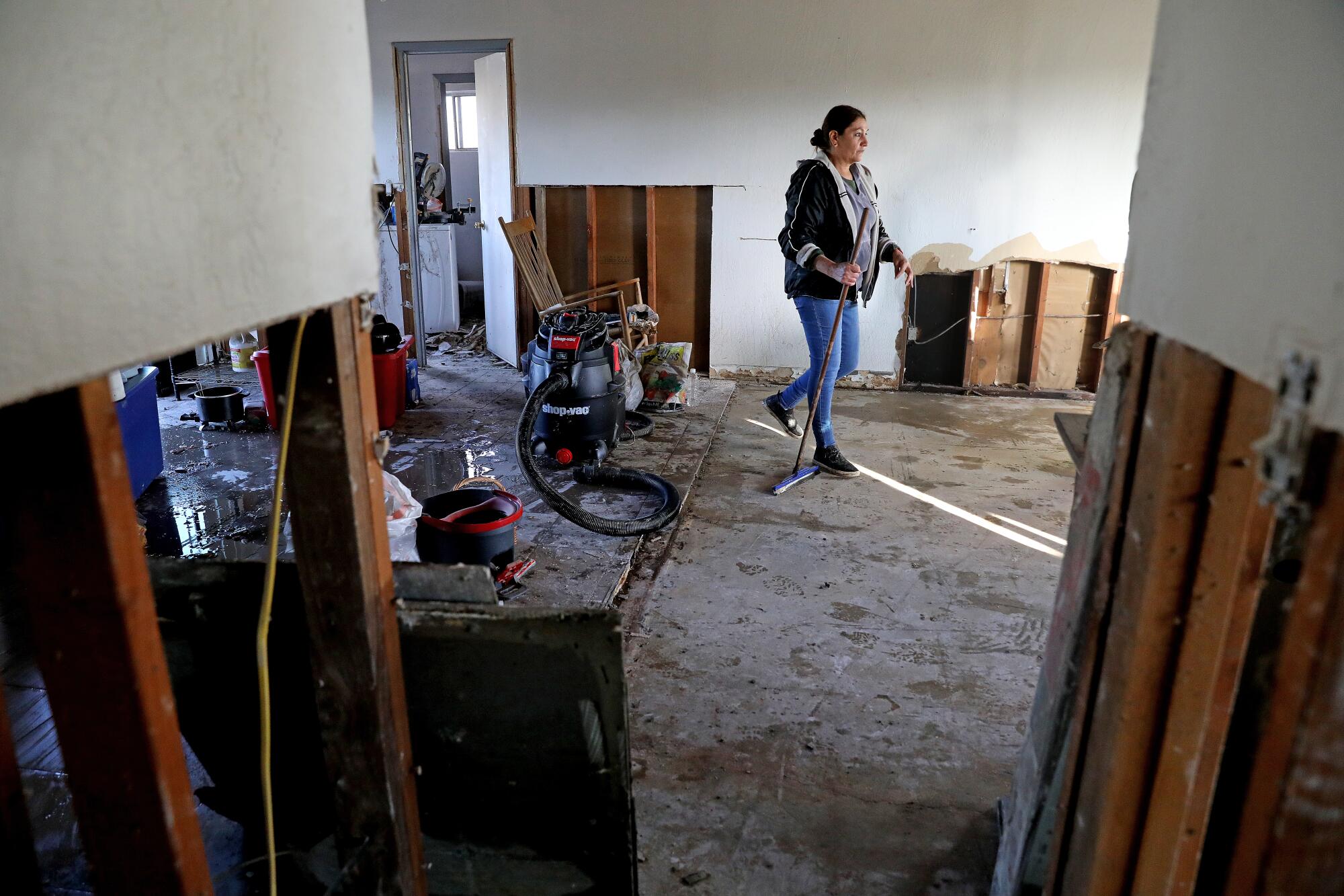 A woman walks across a concrete floor inside a home; sections of drywall have been removed, exposing boards.