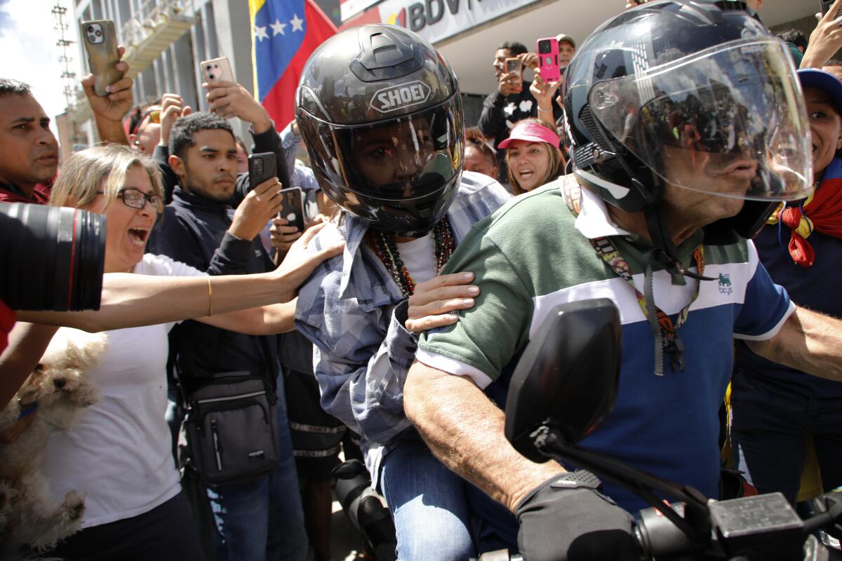 People in a crowd reach out to touch a helmeted woman riding away on the back of a motorbike.