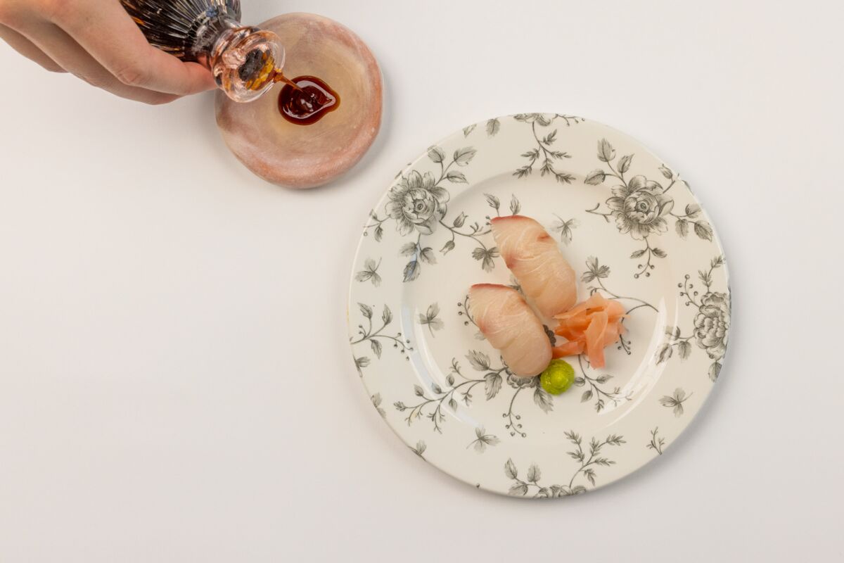 A hand pours soy sauce into a saucer next to a floral-patterned plate of nigiri.