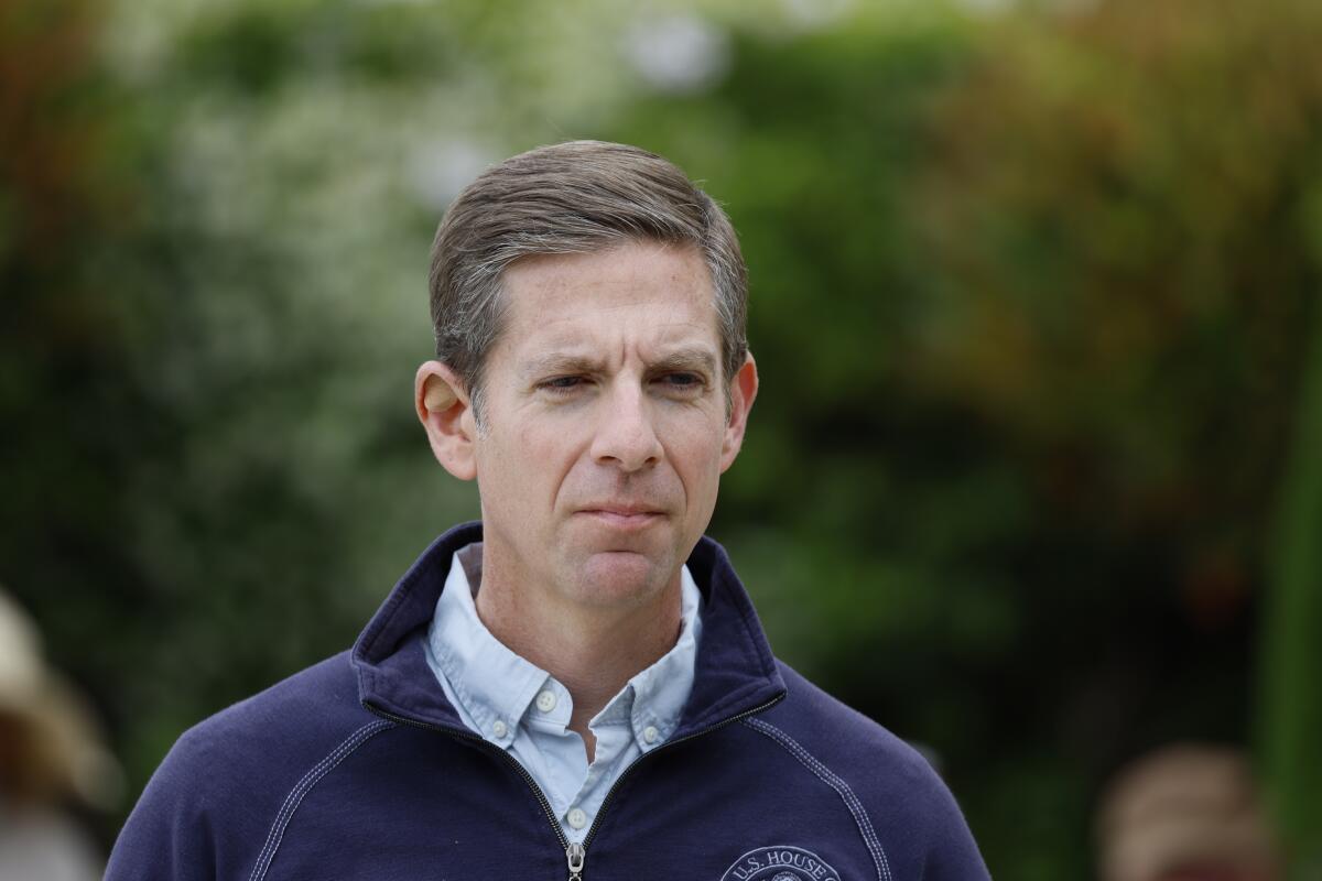Rep.  Mike Levin picture from the shoulders up in front of green leaves, in a blue sweater zippered over a collared shirt