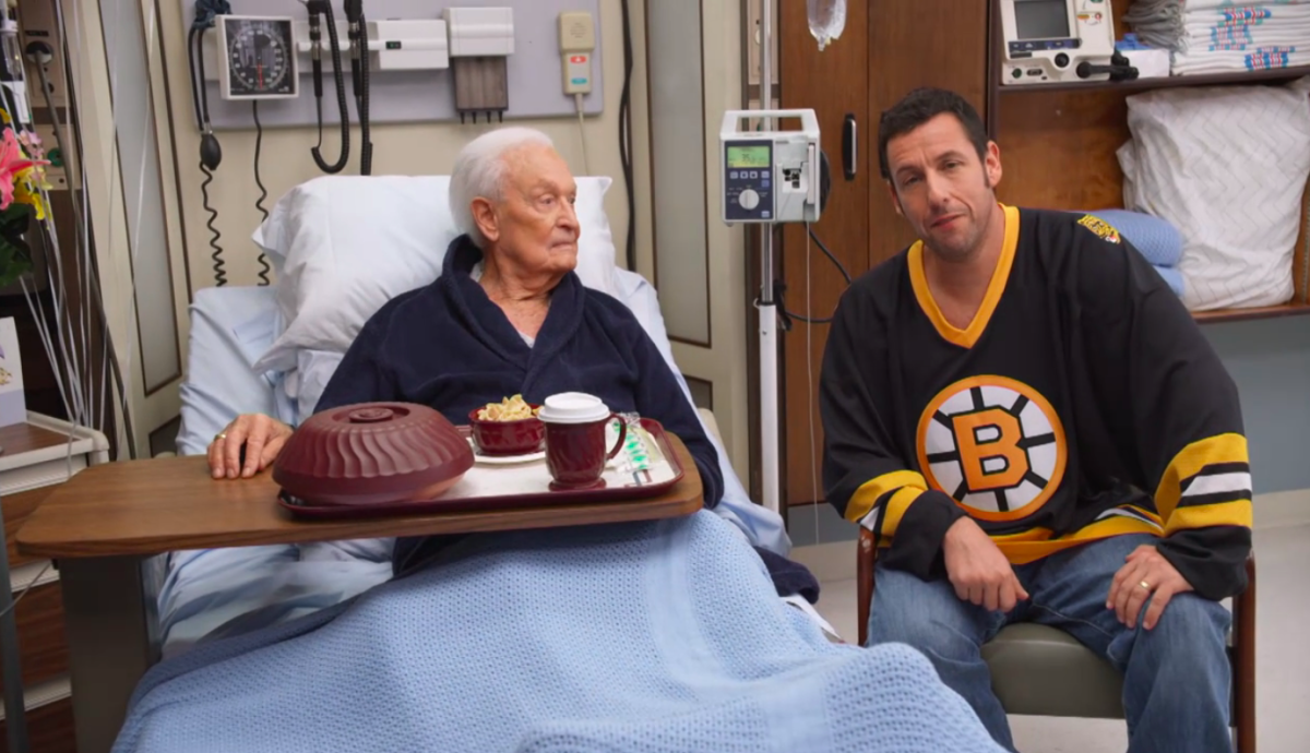 A man in a hockey jersey sits next to an older man in a hospital bed.