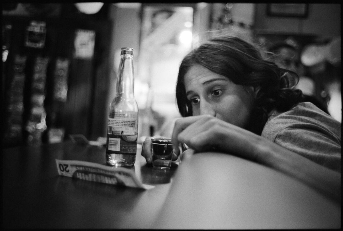 A woman leans her chin on a bar in a black-and-white photo, contemplating a beer and a shot glass.