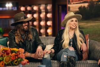 Billy Ray Cyrus with long hair wearing a wide-brimmed hat and jeans sitting next to a blonde woman in a similar outfit