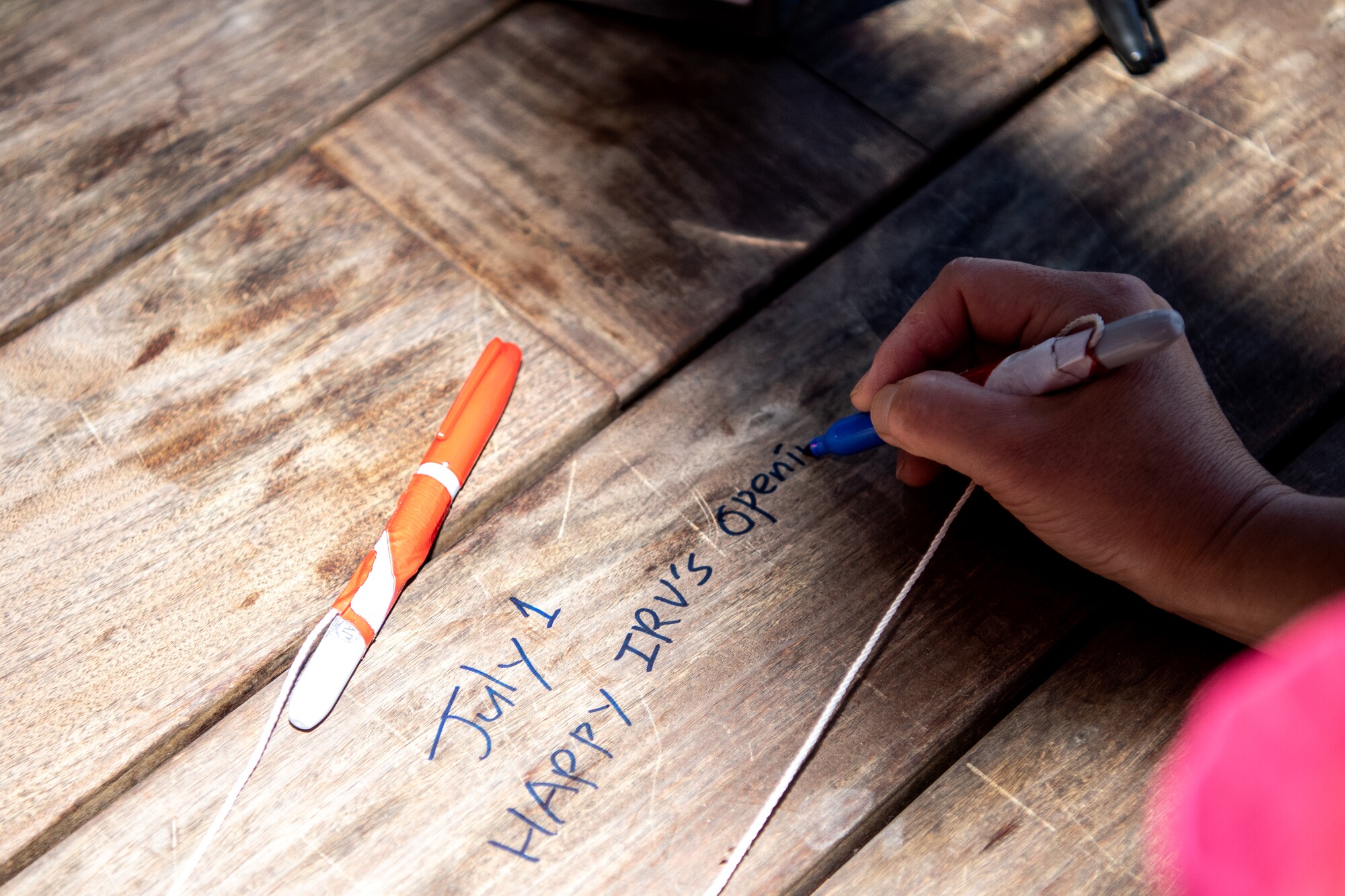 A photo of a hand writing "July 1 Happy Irv's Opening" on a wooden table.