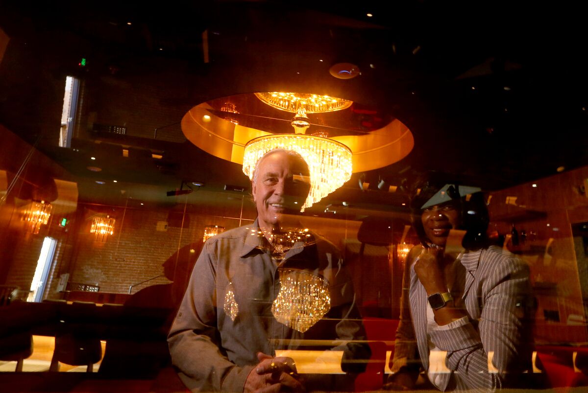 A man and woman pose behind reflective glass with a lighted chandelier in the background