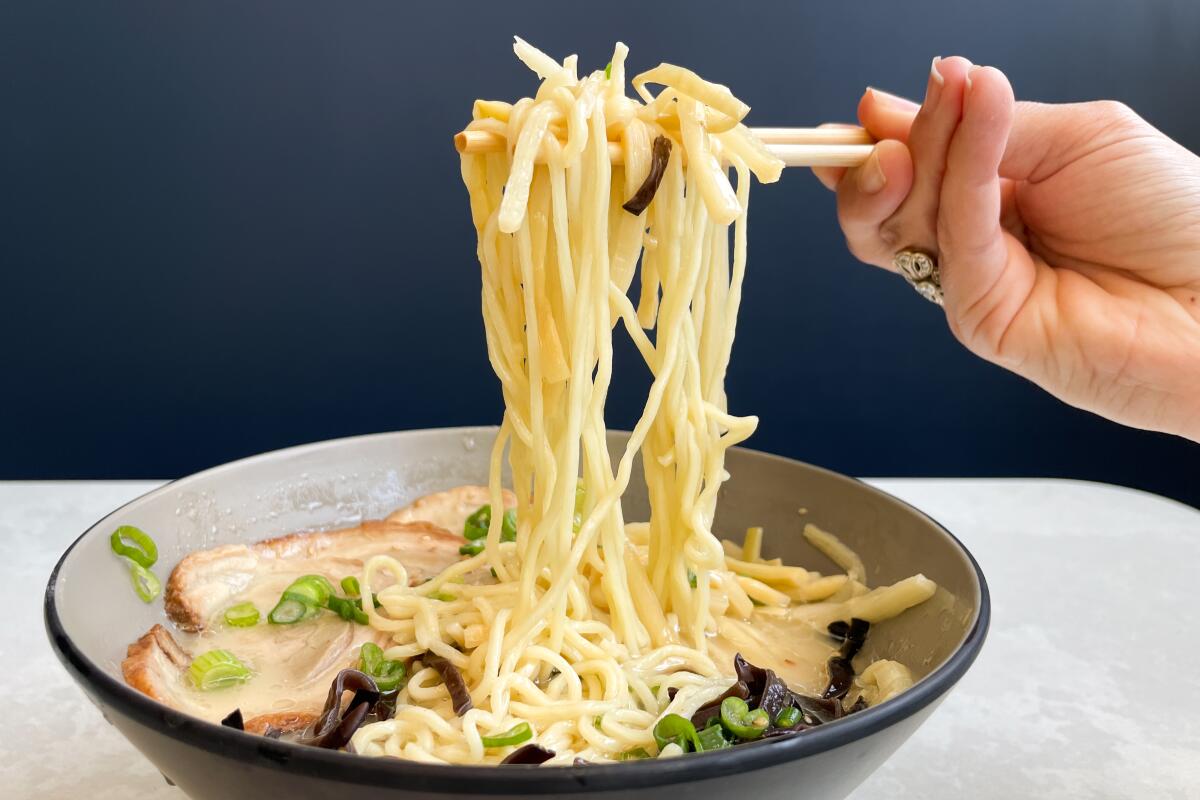 A hand uses chopsticks to hold up ramen from a large bowl