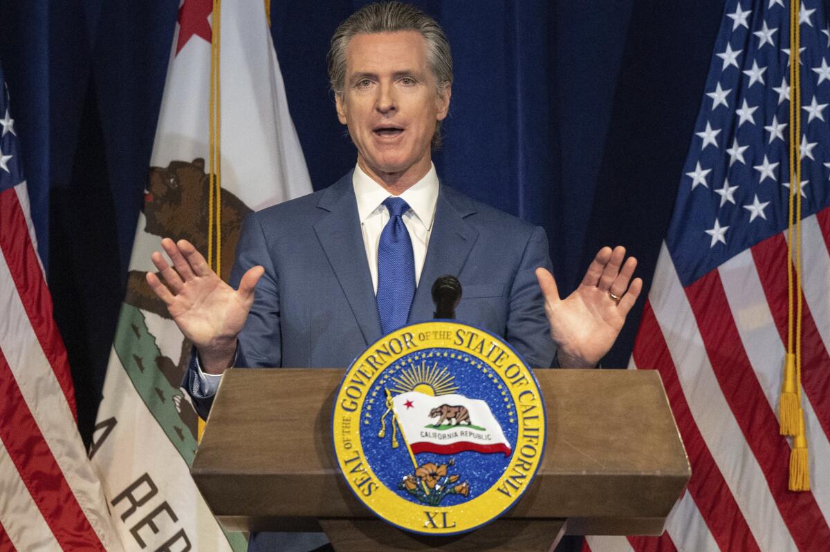 California Gov. Gavin Newsom stands at a lectern and flags are behind him as he holds up his hands to gesture.