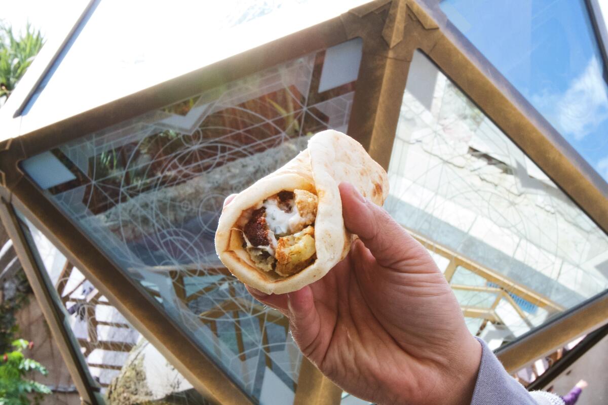 A hand holds a falafel and cauliflower shawarma up against a large mirrored and gold prism in Avengers Campus.