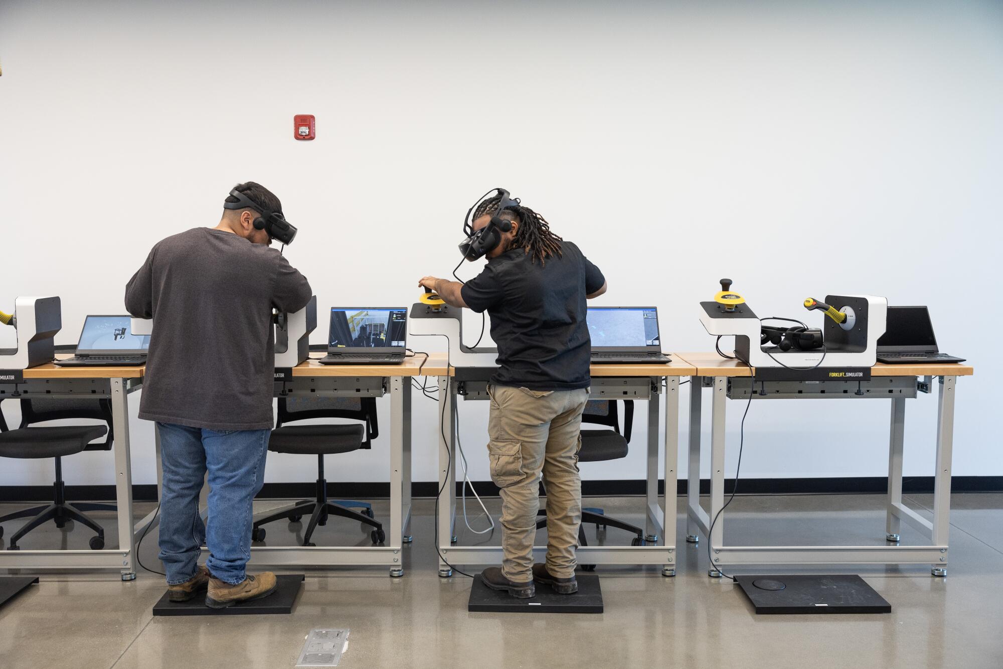 Two men wear virtual reality headsets that simulate forklift operations.  