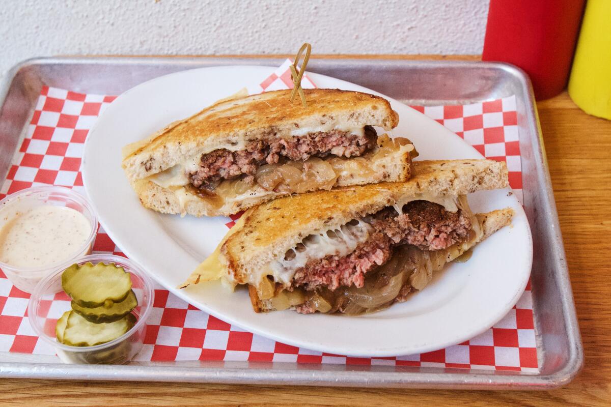 Two halves of a Cassell's  patty melt on rye, Swiss cheese gooey, atop red-and-white checkered paper on a silver tray