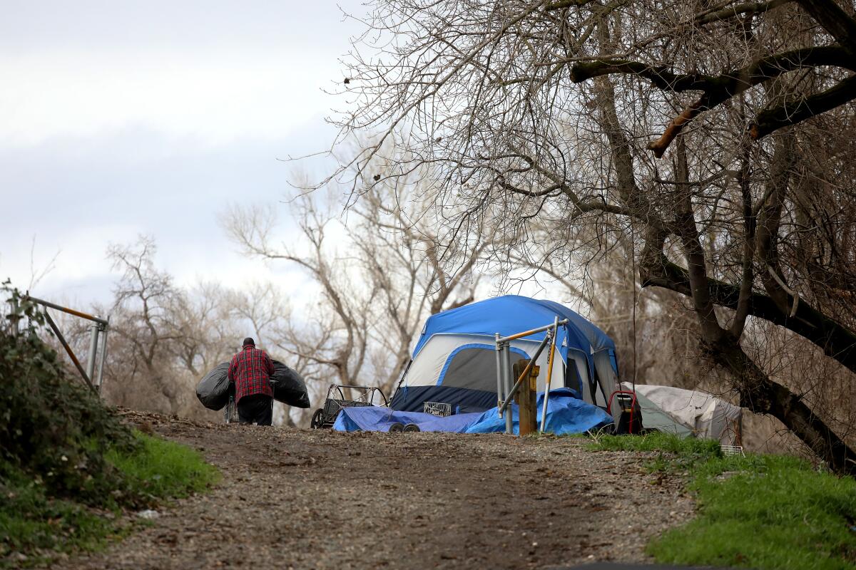 A man pushes a cart filled with belongings next to tents on a dirt trail