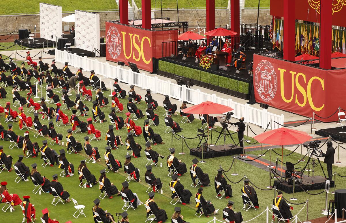 People wearing graduation caps and gowns are seated on a lawn at a commencement ceremony. 