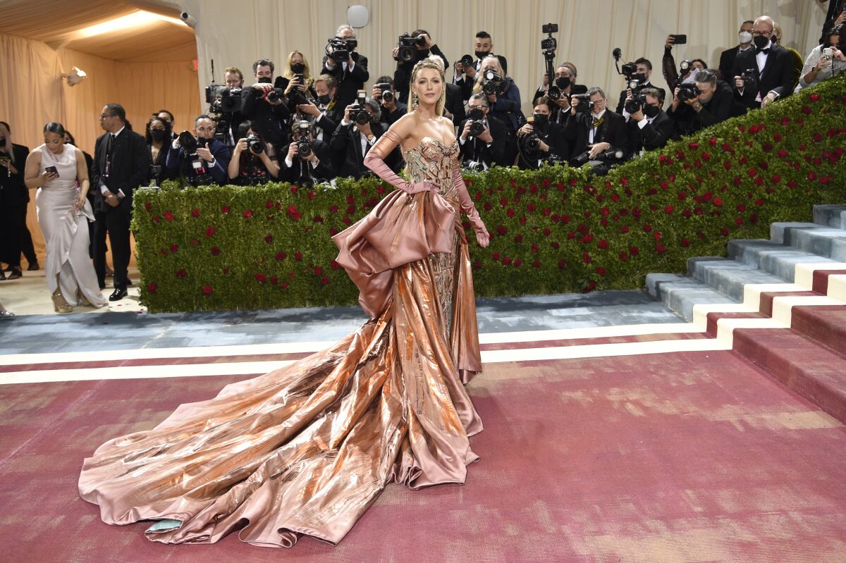 A woman in a strapless dress with a long train poses on a rose-colored carpet for a bank of photographers.