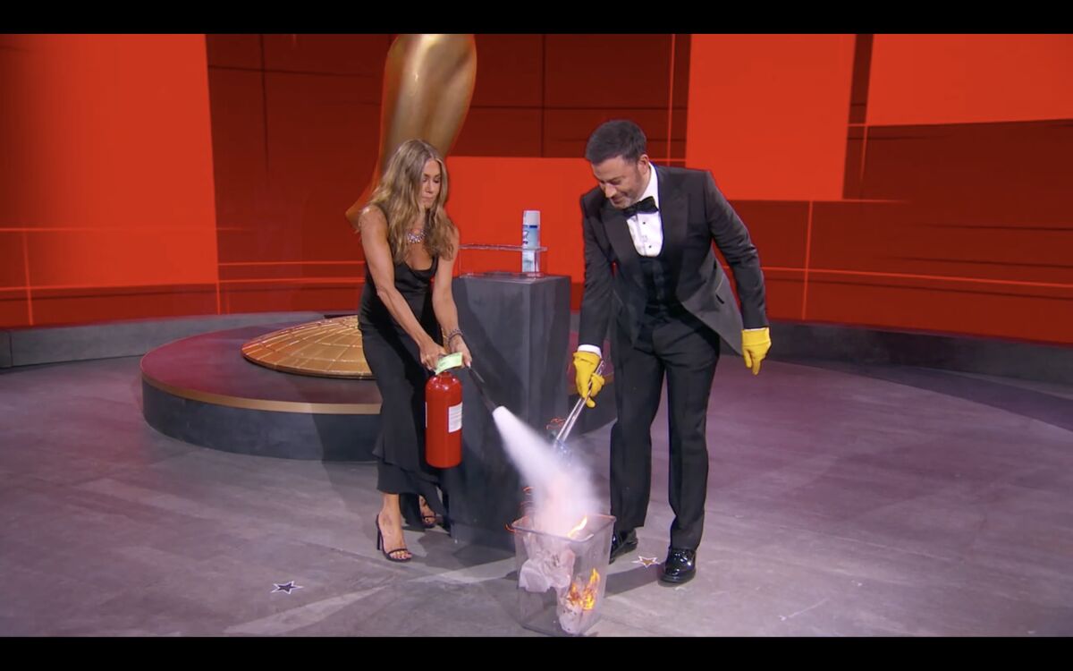 Jennifer Aniston douses a trash can fire at the 2020 Emmys as Jimmy Kimmel uses tongs to grab an envelope from the flames.