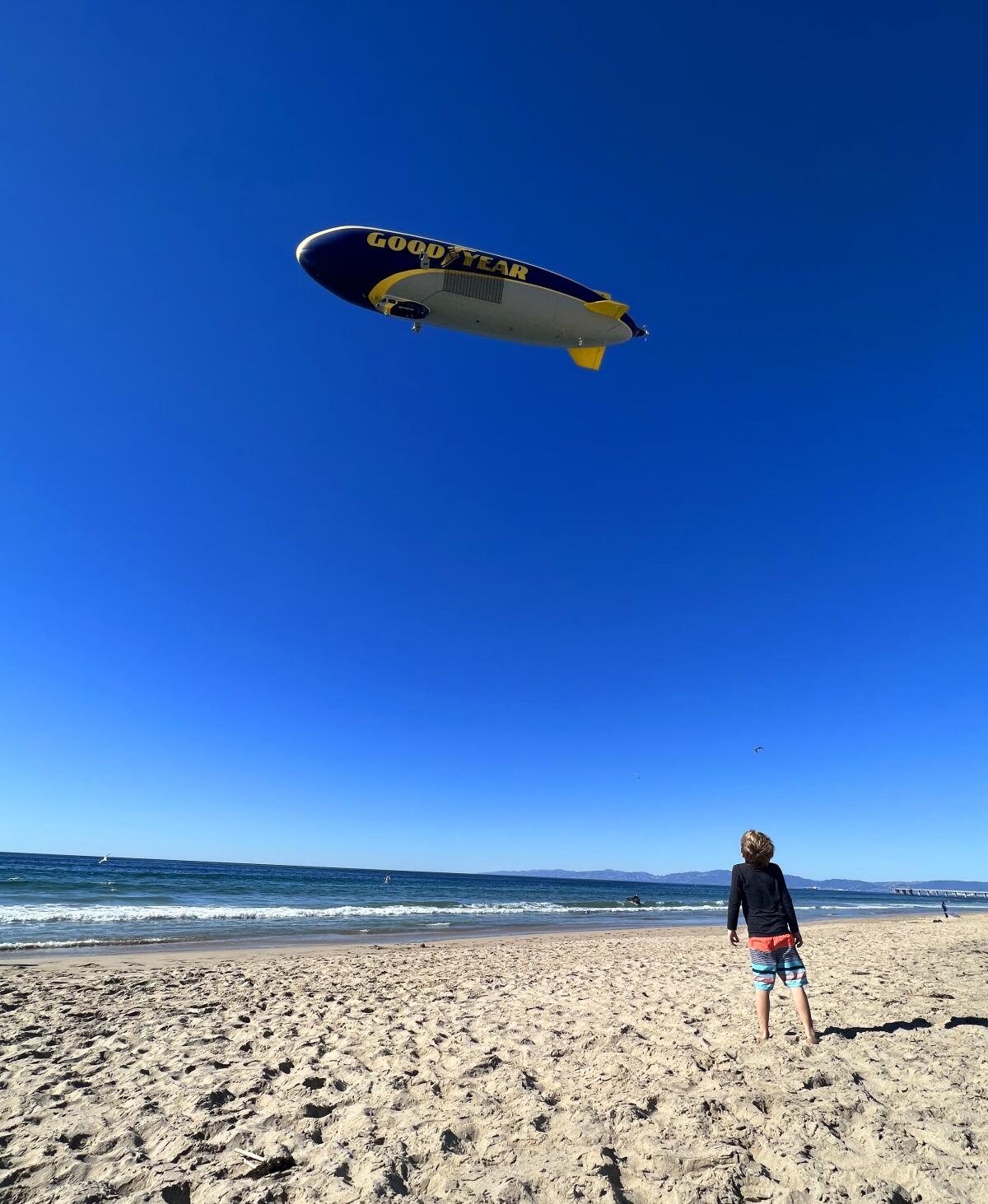 A boy stands on a beach and looks up at a large blimp flying overhead.