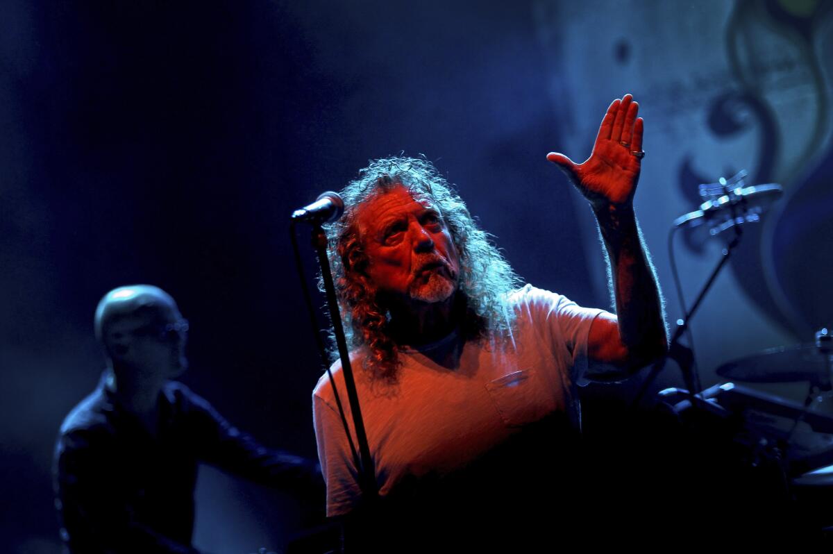Robert Plant wears a gray tee and holds his hand to the sky behind a microphone.
