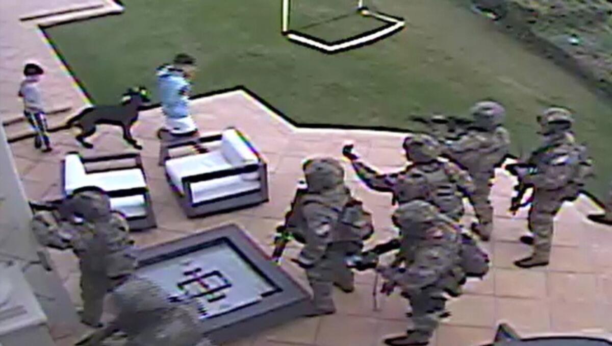 Heavily armed men stand on a patio near two children and a dog.