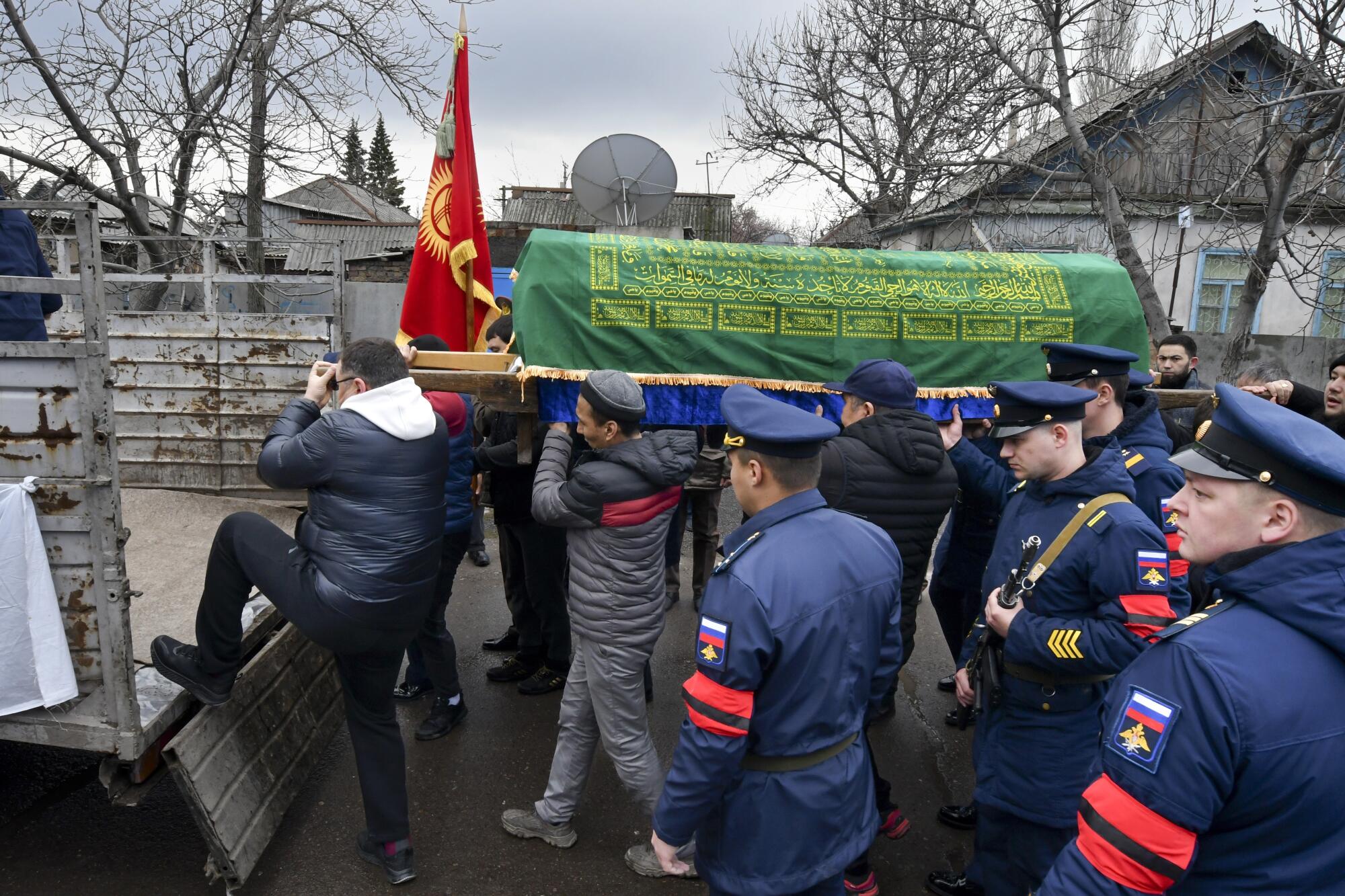 Russian servicemen in blue uniform and hats stand near people loading a coffin, draped in a green cloth, onto a truck. 