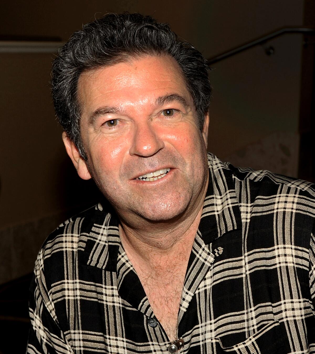 A middle-aged white man with dark brown hair wearing a black and tan plaid shirt with an open collar