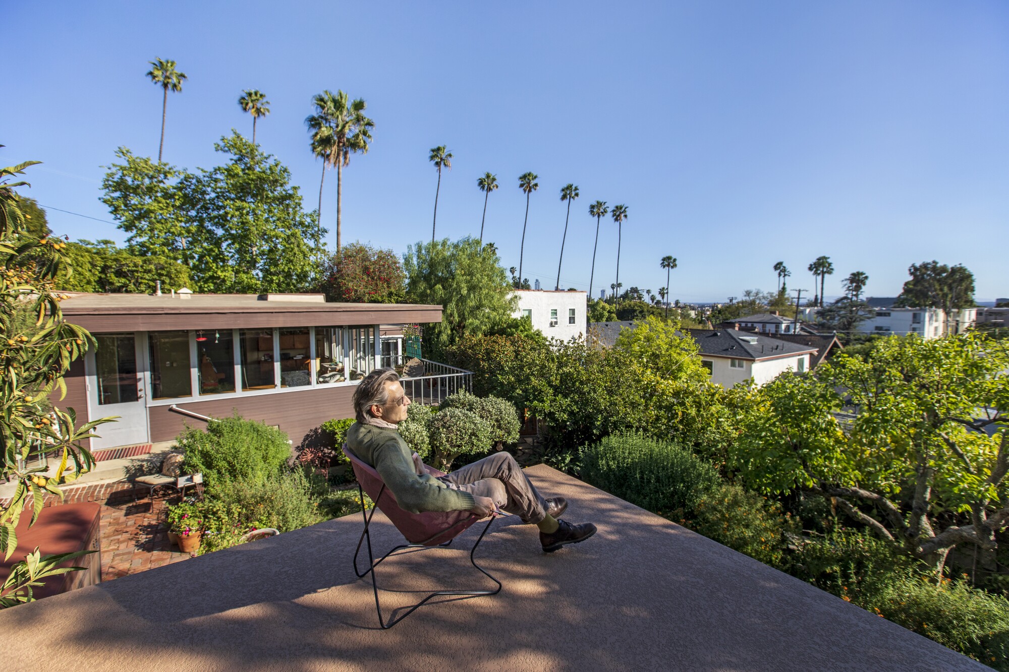 A man sits on a rooftop overlooking palm trees and houses in the distance.