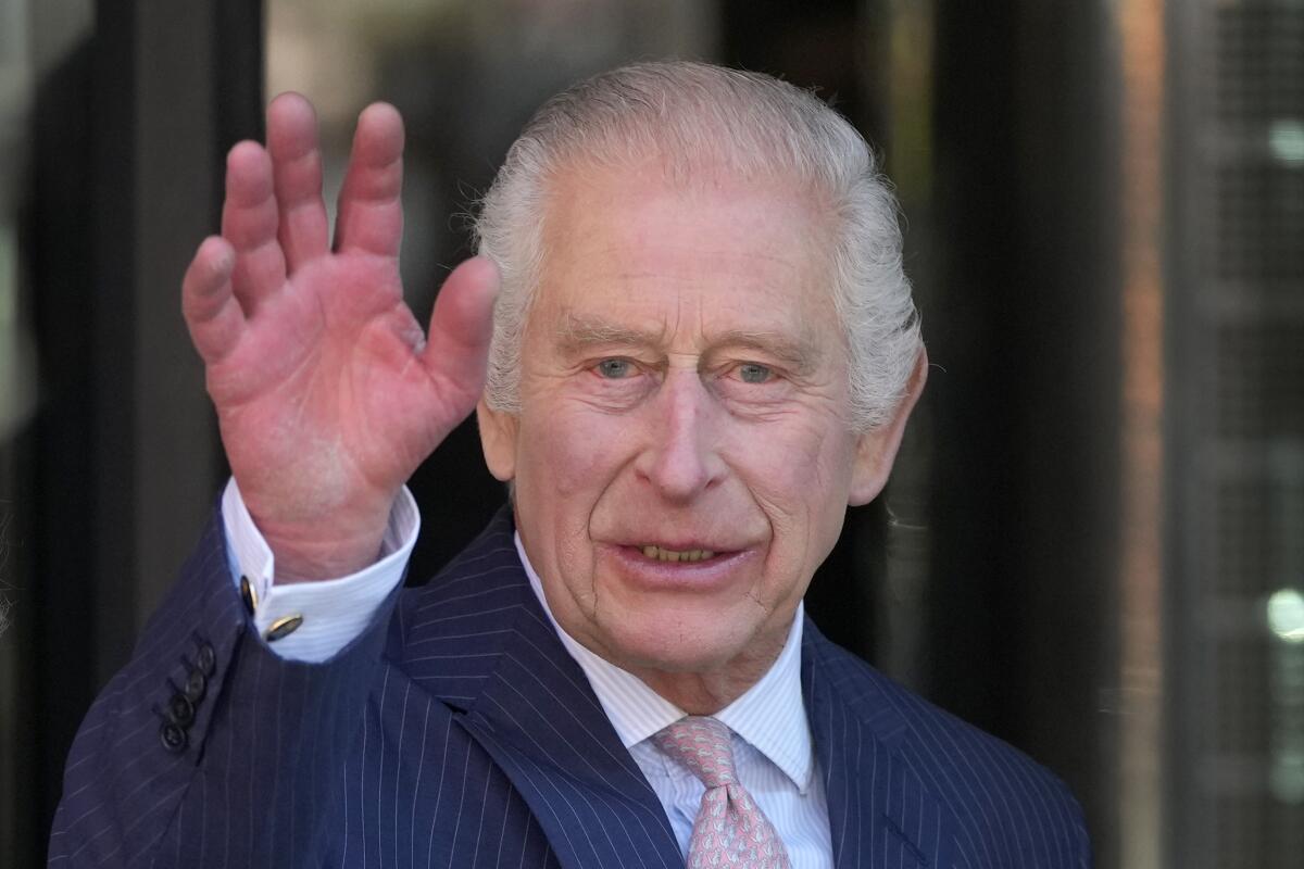 Britain's King Charles III wearing a blue suit and pink tie and waving with one hand