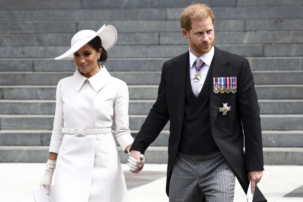 A brunette woman in a white hat and coat holds hands and descends stairs with a red-haired man in a black suit.