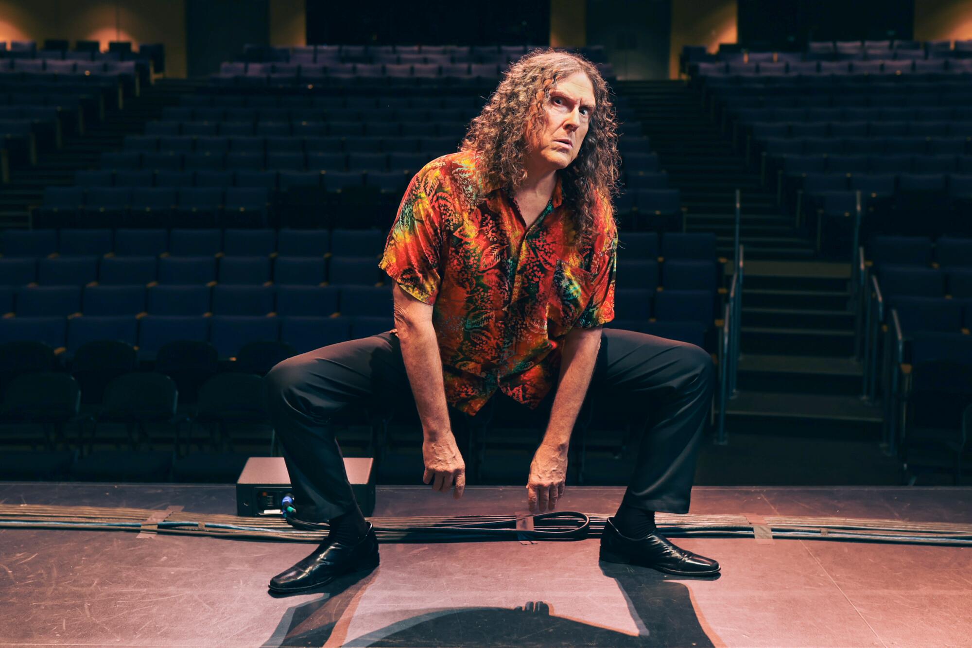 A man squatting on a stage and staring into the camera