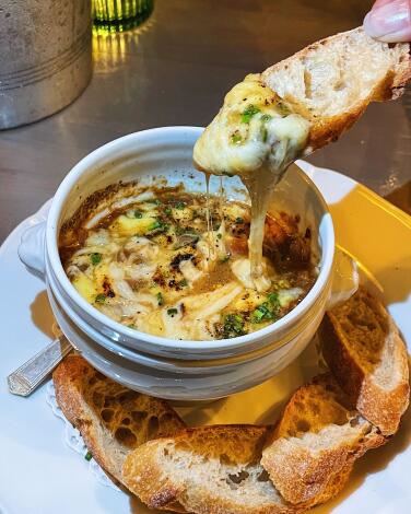 A hand dips bread into a white bowl of cheesy French onion soup dip