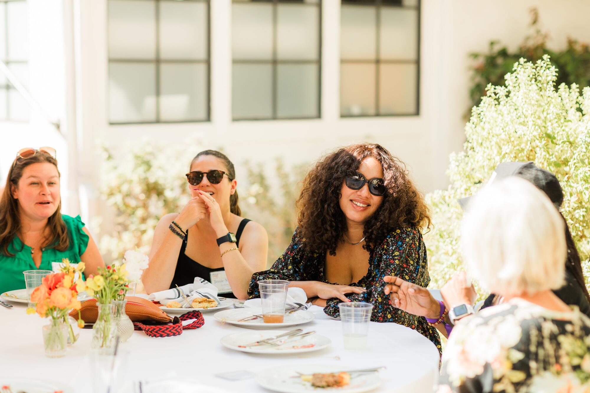 Women seated at an outdoor table smile and talk. 