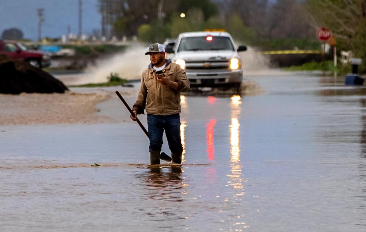A man carrying a long handled implement walks in brown water up to his shins. Behind, a truck throws up a spray of water.