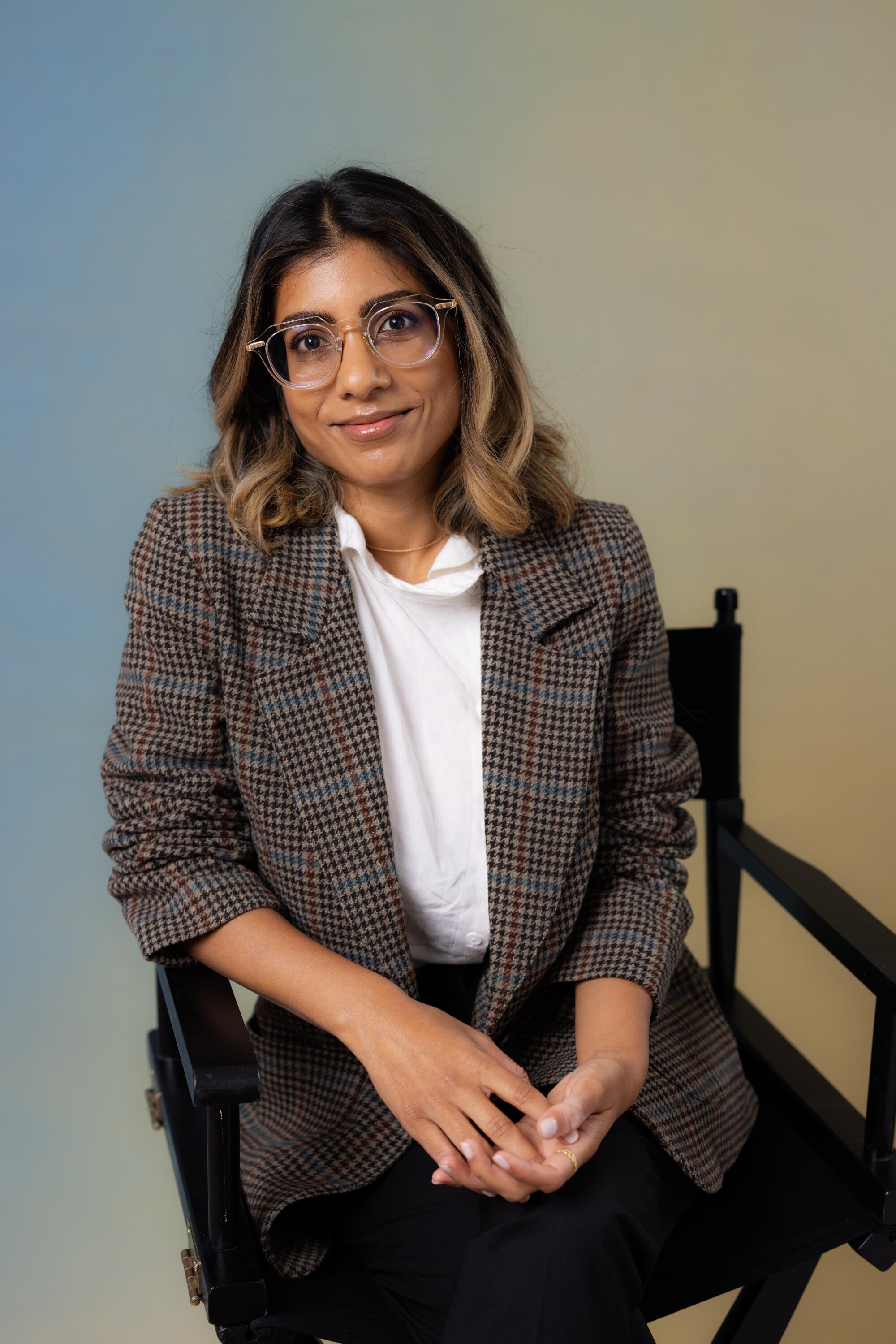 Minhal Baig sits in a chair while wearing a blouse and checked jacket.