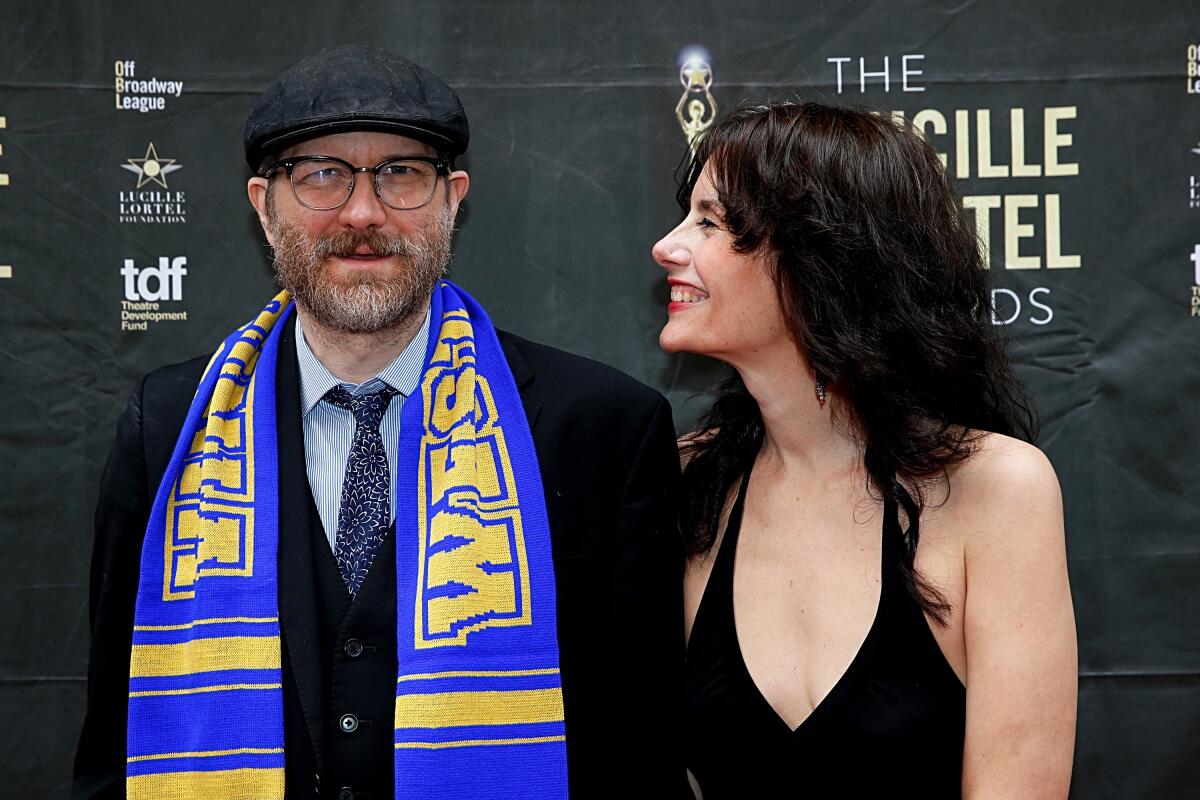 Erik Jensen smiles in a dark suit and hat and a bright scarf as wife Jessica Blank, in a black dress, smiles at him