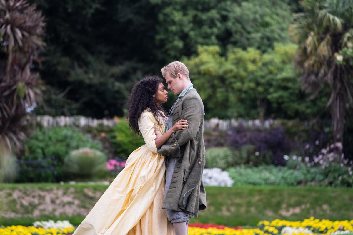 A woman in a yellow dress and a man in a grey coat embrace in a garden.