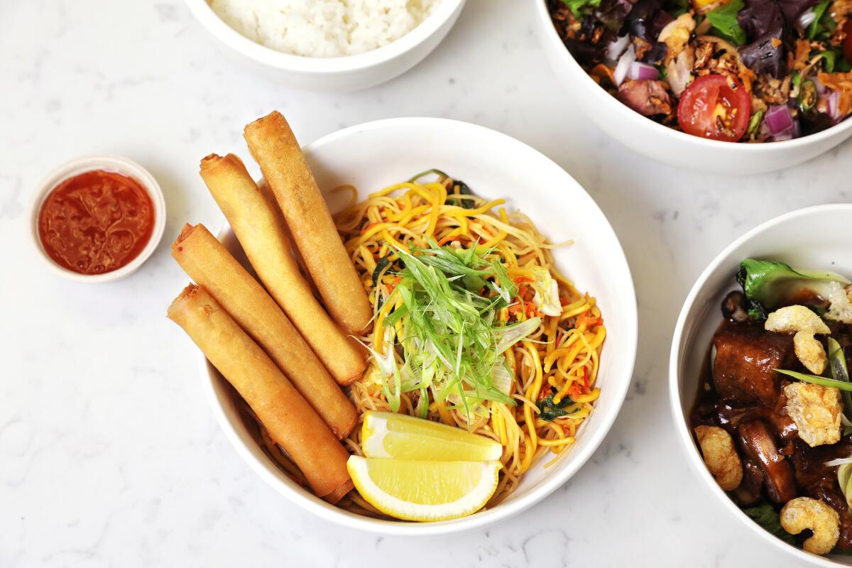 An overhead photo of baboy tim, sisig salad, white rice, pancit and lumpia from Petite Peso, photographed on white marble.