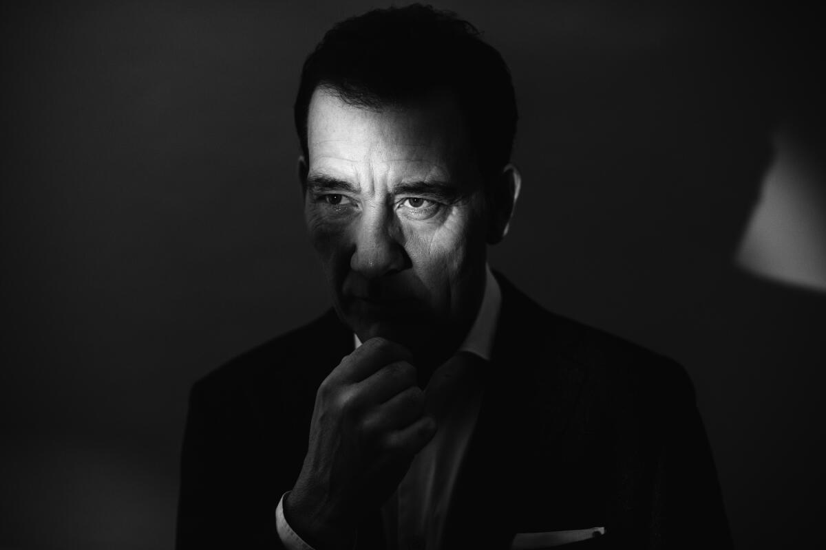 Clive Owen with his hands on his chin in a dark room with a ray of light across his face.