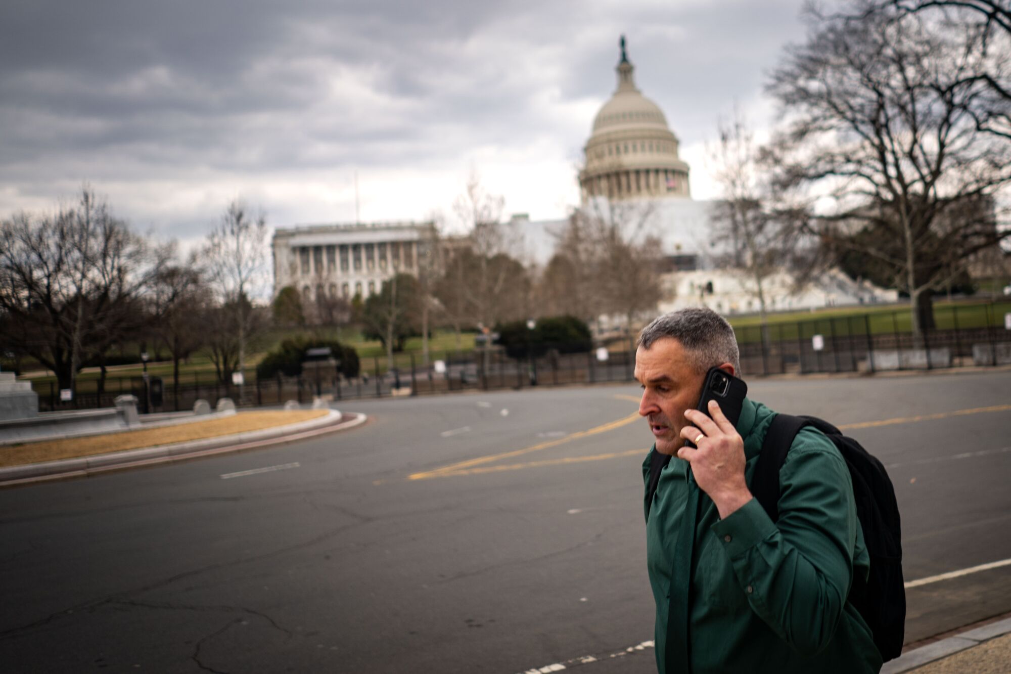 A man holds a cellphone to his ear as he walks along a sidewalk with the U.S. Capitol visible in the background.
