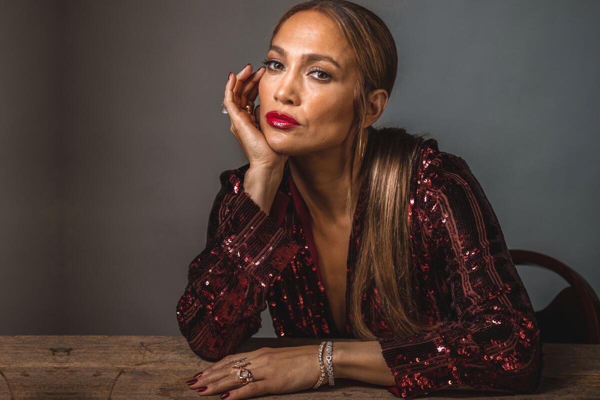Jennifer Lopez leaning her chin on one hand while resting the other on a table she is sitting behind