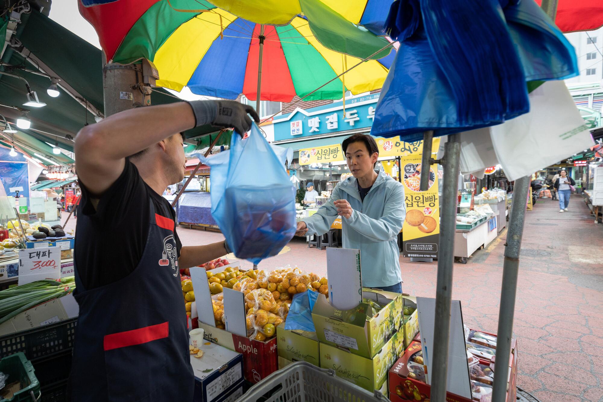 One man holds up a bag of food while another waits under a brightly colored umbrella